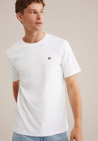 Mlad moški gleda na stran, oblečen v enostavno belo majico s kratkimi rokavi, na prsih ima majhen črn logotip, ter svetlo modre kavbojke.