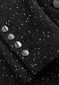 Gros plan sur un tissu en tweed noir avec des éclats blancs et cinq gros boutons ronds argentés brillants alignés en série.
