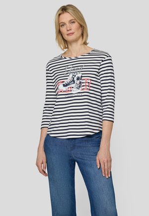 Frau mit blondem Haar, die ein marineblau-weiß gestreiftes Shirt mit Sneaker-Grafik und dem Schriftzug "Good Vibes" trägt, kombiniert mit blauen Jeans.