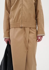 Veste beige légère avec taille élastique, manches longues et texture lisse. Assortie à une jupe ample et un sac noir.