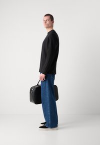 Camisa negra de manga larga, jeans anchos azul, zapatos de ante negros, sosteniendo un bolso rectangular de cuero negro con un asa superior.