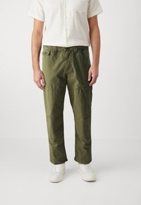 Green cargo pants featuring a relaxed fit, multiple pockets, and a smooth texture. Paired with white sneakers and a short-sleeve shirt.