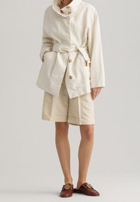 Giacca belted in cotone color beige chiaro con bottoni dorati, abbinata a pantaloncini color crema e mocassini marroni scuri con dettagli dorati.