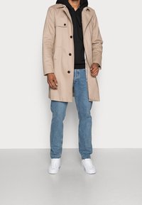 Cappotto beige con design doppiopetto, quattro bottoni frontali e tasche laterali, indossato sopra un hoodie nero, abbinato a jeans blu e sneaker bianche.