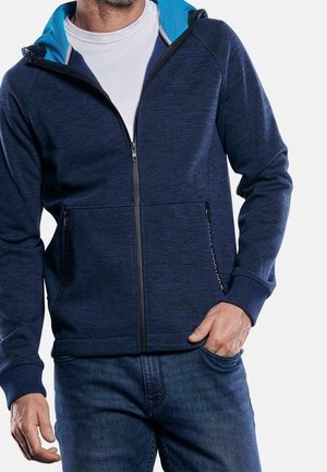 Mann trägt einen dunkelblauen Kapuzenpulli mit Reißverschluss und hellblauer Kapuzenfutterung über weißem T-Shirt und blauen Jeans, eine Hand in der Tasche.