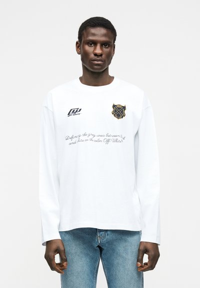 Άνδρας που φοράει λευκό μακρυμάνικο πουκάμισο με μαύρα και χρυσά λογότυπα και γράμματα, σε συνδυασμό με ανοιχτό μπλε τζιν, στέκεται μπροστά σε απλό φόντο.