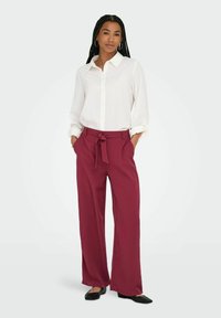 Chemise blanche à boutons avec manches retroussées, associée à un pantalon bordeaux à jambes larges avec une ceinture à nouer. Des ballerines noires complètent la tenue.