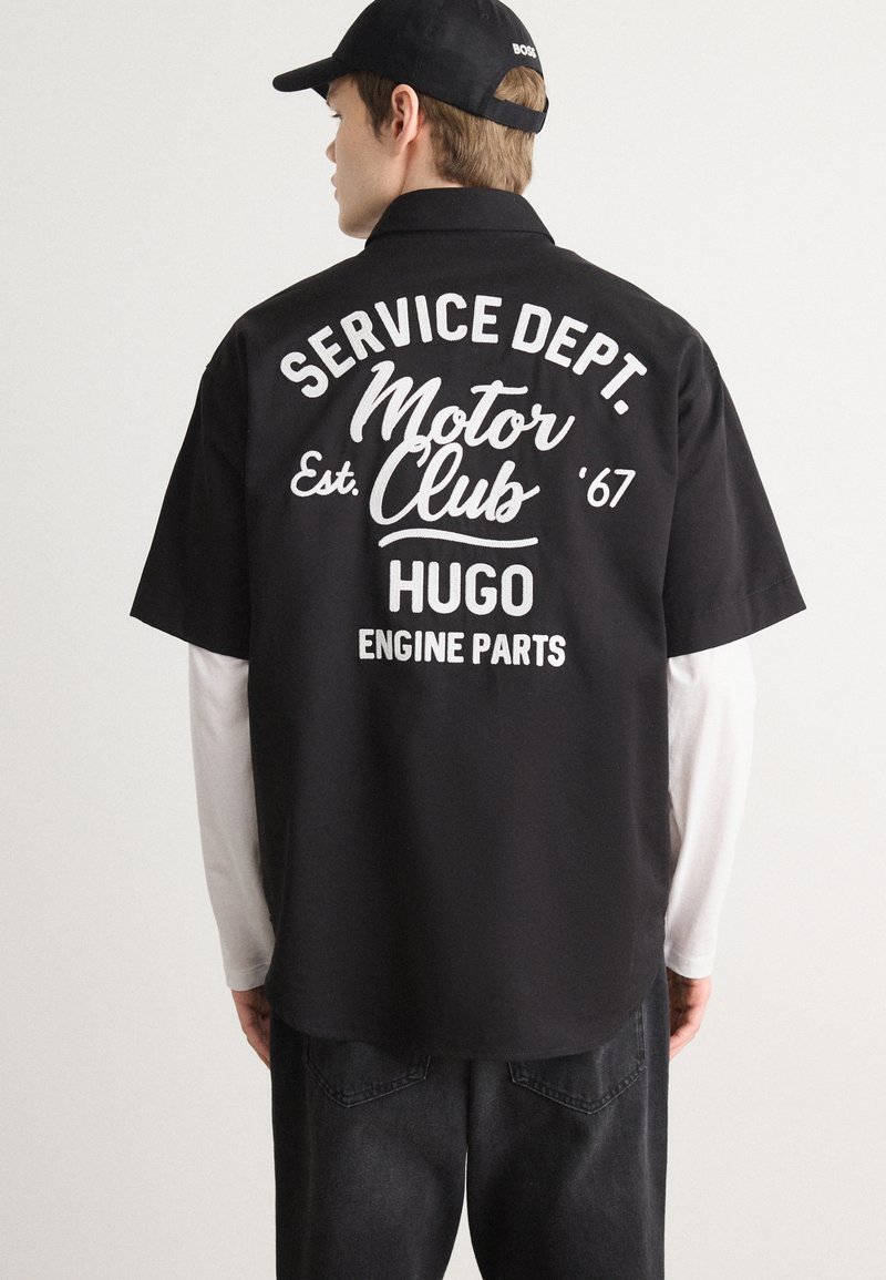 Zwart shirt met korte mouwen met witte tekst op de achterkant; leest "SERVICE DEPT. Motor Club Opgericht '67 HUGO MOTORDELEN." Casual ontwerp.