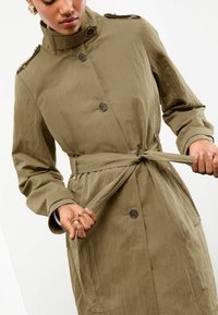 Persoon die de riem van een khaki trenchcoat met knopen, schouderepauletten en een hoge kraag strijkt.