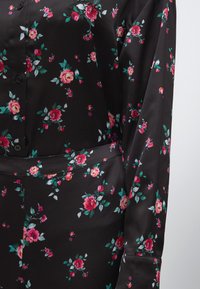 Blouse noire à motifs floraux, manches courtes, devant boutonné, avec un motif de roses roses et de feuilles vertes sur un tissu lisse.