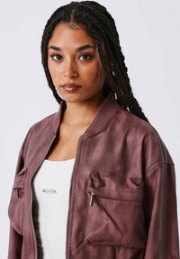 Bordeaux bomberjack met twee voorzakken, ritssluiting, ribgebreide kraag en gestructureerde stof. G gedragen over een witte tanktop.