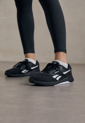 Zwarte sport schoenen met een gestructureerde mesh bovenkant, witte accenten en een gewatteerde zool. Draagt in combinatie met zwarte leggings, wat een slank design benadrukt.
