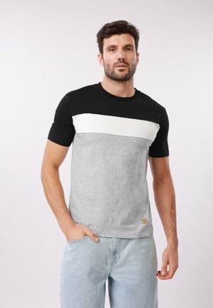 Homme portant un T-shirt à blocs de couleurs noir, blanc et gris avec un jean bleu clair, debout avec une main dans la poche devant un fond blanc.