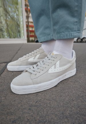 Lys grå ruskinds sneakers med hvide detaljer, tekstureret sål og flade snørebånd. Synlig syning og perforerede detaljer på siden.