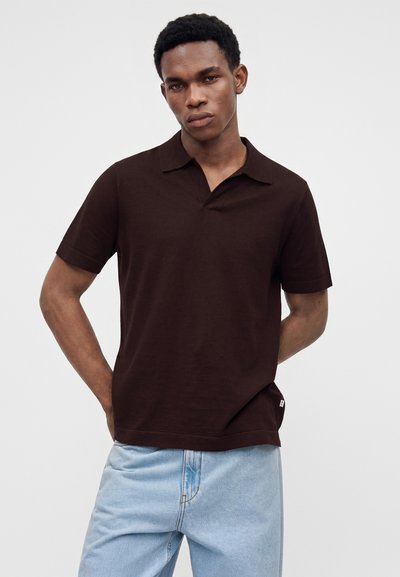 Jeune homme portant un polo marron foncé à manches courtes et un jean bleu clair, debout avec les mains dans le dos.