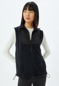 Gilet in pile nero con zip frontale, presenta un pannello nero liscio sul petto, tasche laterali e cordini regolabili all'orlo.