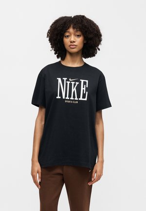 Kvinde med krøllet hår iført en sort Nike Sports Club T-shirt og brune bukser, stående foran en ensfarvet lys baggrund.