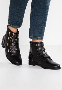 Bottes noires à la cheville en cuir avec trois sangles à boucle et des clous dorés, présentant un petit talon et un détail de fermeture éclair sur le côté.