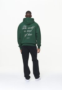 Felpa verde con testo bianco sulla schiena che recita "Il mondo è stanco dell'odio", abbinata a pantaloni neri e scarpe bianche. Design semplice.