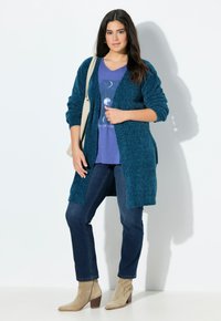 Mörkgrön stickad cardigan över en lila grafisk T-shirt, i kombination med mörka jeans och beige ankelstövlar. Neutral bakgrund.
