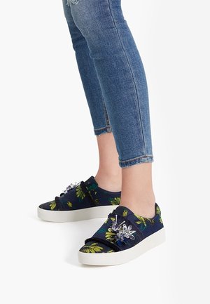 Personne portant des baskets à enfiler bleues à motif floral avec des ornements décoratifs et un jean skinny bleu, sur fond blanc.