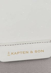 Vitt och grått Kapten & Son tygprodukt med en guldlogotyp. Slät textur med subtila prickar och kontrasterande sömnadsdetaljer.