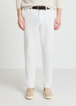 Persona con pantaloni bianchi, maglione beige, cintura intrecciata scura e scarpe beige senza lacci, in piedi davanti a uno sfondo chiaro e semplice.
