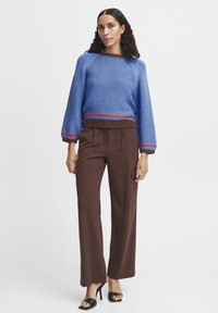 Blaues Strickpullover mit einem gerippten braunen Saum und roten Akzenten, kombiniert mit hochgeschnittenen braunen Hosen mit Seitentaschen und geradem Schnitt.