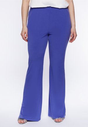 Kobieta w jasnoniebieskich szerokich spodniach z wysokim stanem z małymi rozcięciami po bokach, w połączeniu z sandałami na palce zdobionymi ćwiekami.