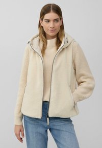 Frau in einem cremefarbenen Strickpullover und einer gefütterten Fleece-Weste mit Kapuze, kombiniert mit blauer Jeans, die vor einem schlichten hellen Hintergrund steht.