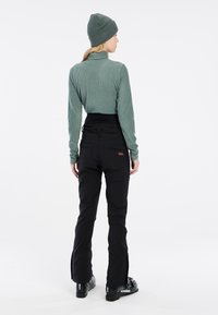 Col roulé côtelé vert, pantalon de ski imperméable noir avec poches arrière, et bottes de ski noires avec logos blancs. Design simple, fonctionnalité pratique.