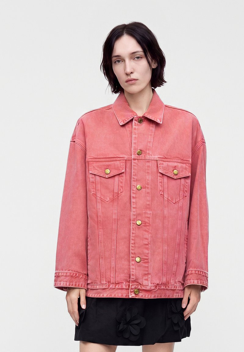 Jeune personne aux cheveux courts et foncés portant une veste en jean rouge délavée et oversize ainsi qu'une jupe noire avec des appliques florales, debout sur un fond uni.