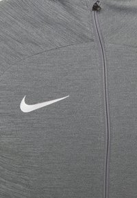 Grijze Nike jas met ritssluiting, gemaakt van gestructureerde stof, met een wit swoosh-logo en een rits aan de voorkant.