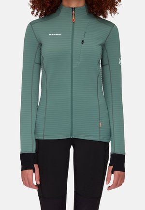 Groene fleecejas met rits, borstzak, duimgaten en Mammut-logo, gedragen over een zwarte broek.