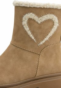 Botín de ante color topo con un cuello forrado de piel y un recorte en forma de corazón relleno de piel texturizada. Presenta detalles de costura y una suela de goma.