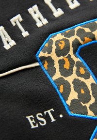 Svart tyg med upphöjd vit broderad text, en stor leopardmönstrad "C"-applikation kantad med blå sömnad, samt "EST." i vitt.
