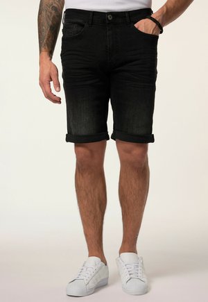 Mann trägt schwarze hochgekrempelte Jeans-Shorts, weiße Sneaker, ein weißes Hemd, mit einem tätowierten Arm und der Hand in der Tasche.