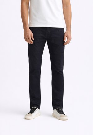 Uomo che indossa pantaloni neri aderenti, maglietta bianca e sneakers bianche con lacci neri, in piedi davanti a uno sfondo bianco.