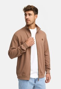 Brown zip-up sweatshirt with a ribbed texture, high collar, and logo patch. Worn over a white t-shirt and paired with light blue jeans.