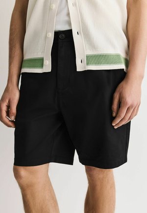 Mann trägt schwarze knielange Shorts und eine weiße ärmellose Strickweste mit grünen Streifen am Saum, steht mit entspannten Händen.