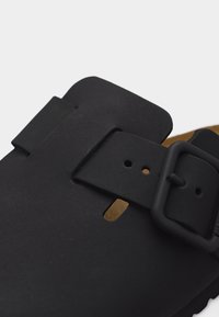 Κοντινό πλάνο μαύρης ματ αγκράφας σε λουρί σε σκούρο καφέ σανδάλι με υφή στη σόλα, που αναδεικνύει τον μηχανισμό στερέωσης.