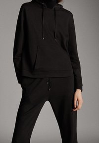 Mustat huppari ja yhteensopivat jogger-housut, jotka on valmistettu pehmeästä ja joustavasta materiaalista. Hupparissa on etutasku ja nyörit; jogger-housuissa on joustavat henkseleitä.