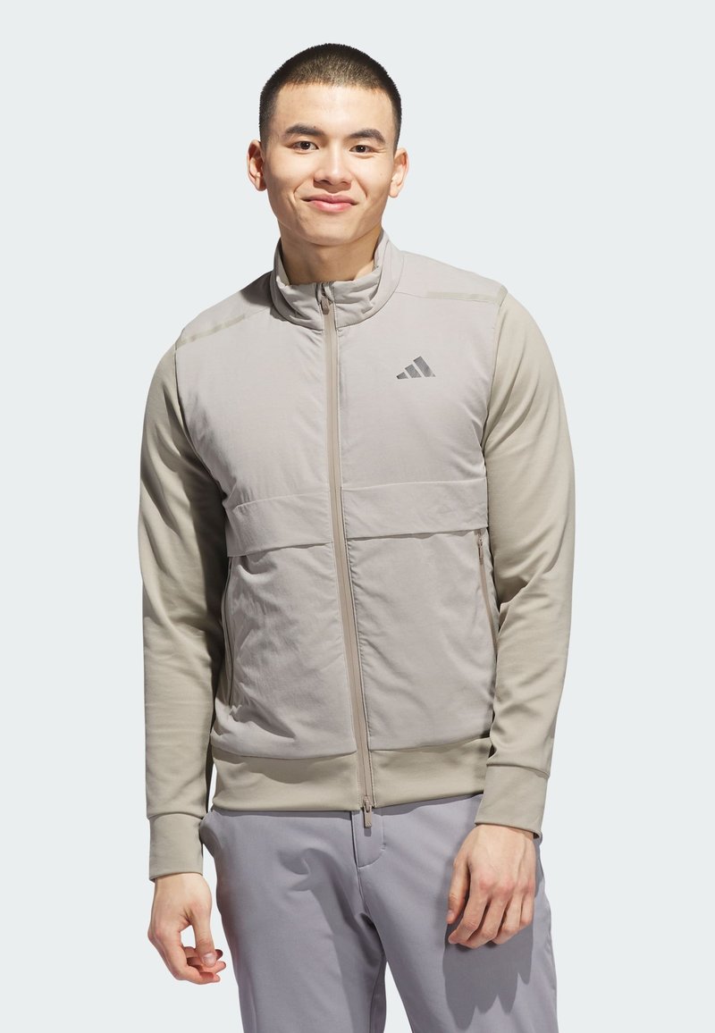 Svijetlo siva zip-jakna s visokim ovratnikom, prednjim džepovima i teksturiranim tkanim panelima; sadrži Adidas logo na gornjem lijevom dijelu prsnog koša.