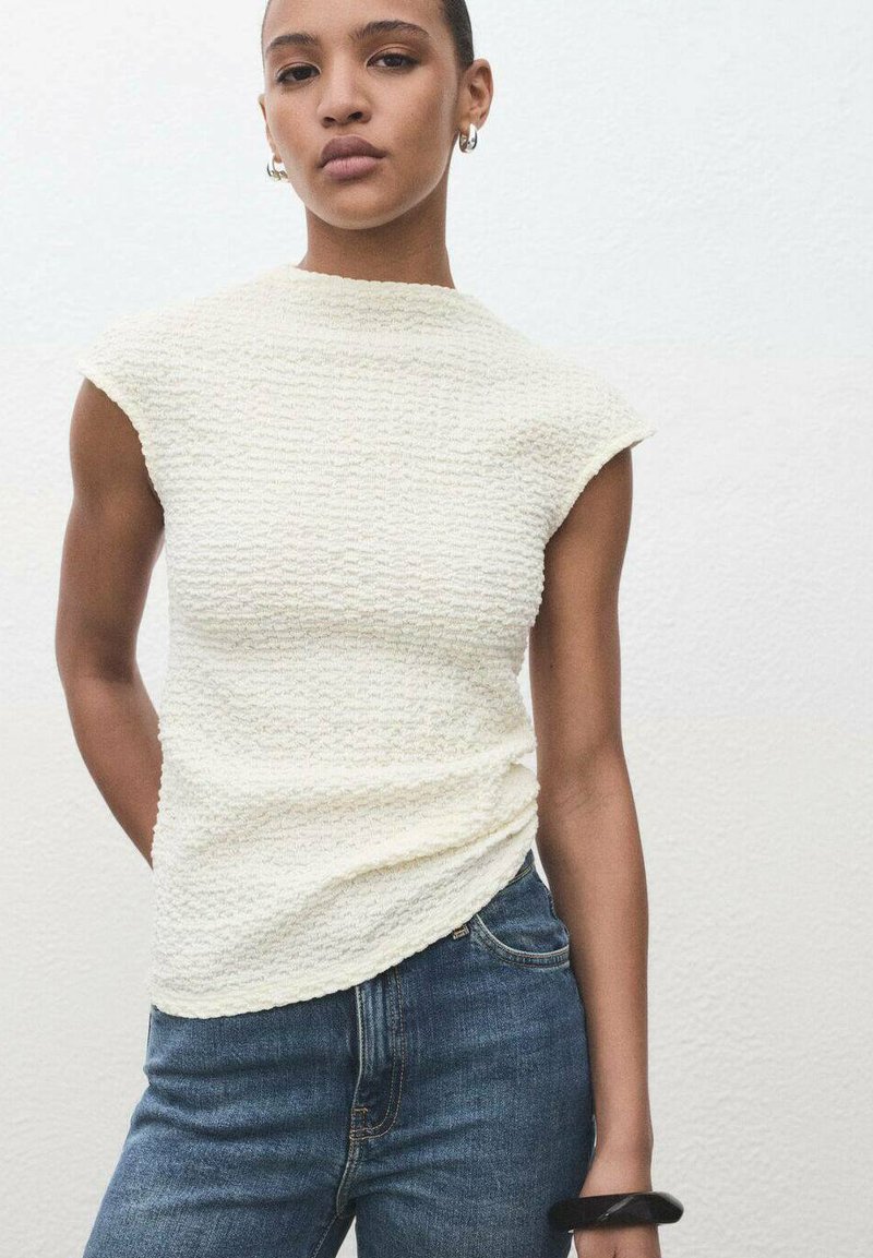Top bianco a collo alto con maniche a cappuccio, abbinato a jeans in denim blu. Il top presenta un motivo ondulato sottile e una silhouette aderente.