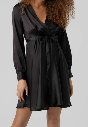 Femme portant une robe portefeuille noire en satin à manches longues, avec un nœud noué à la taille et une jupe évasée, sur un fond uni.
