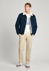 Jachetă de denim bleumarin cu guler alb pufos, purtată peste un hanorac grafic de culoare deschisă, asortată cu pantaloni bej și adidași albi.