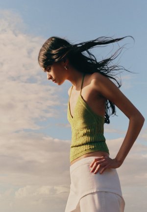 Jonge vrouw in groene mouwloze top en witte rok staat met handen op de heupen, haar waait in de wind tegen een bewolkte lucht.
