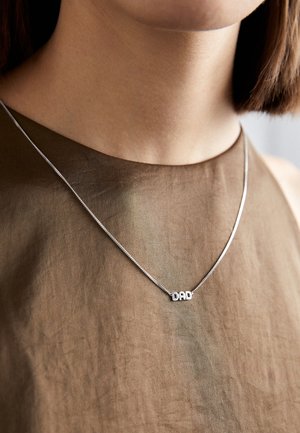 Gros plan d'une personne portant un collier chaîne en argent avec un pendentif portant le mot « DAD » sur un top en tissu marron.