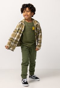 Grünes Langarmshirt mit gelbem Logo, gemustertes olivgrünes und cremefarbenes Flanellhemd, olivgrüne Stretchpants und schwarze Vans-Sneaker.