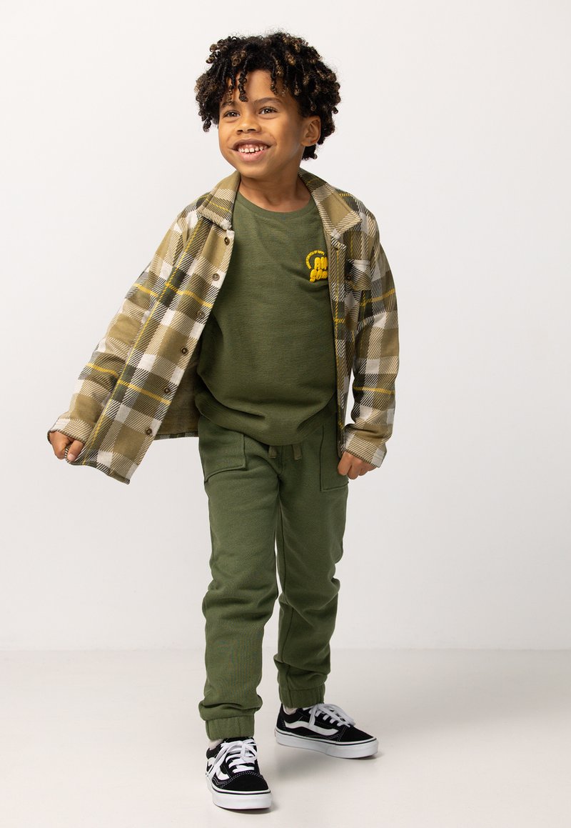 Grünes Langarmshirt mit gelbem Logo, gemustertes olivgrünes und cremefarbenes Flanellhemd, olivgrüne Stretchpants und schwarze Vans-Sneaker.